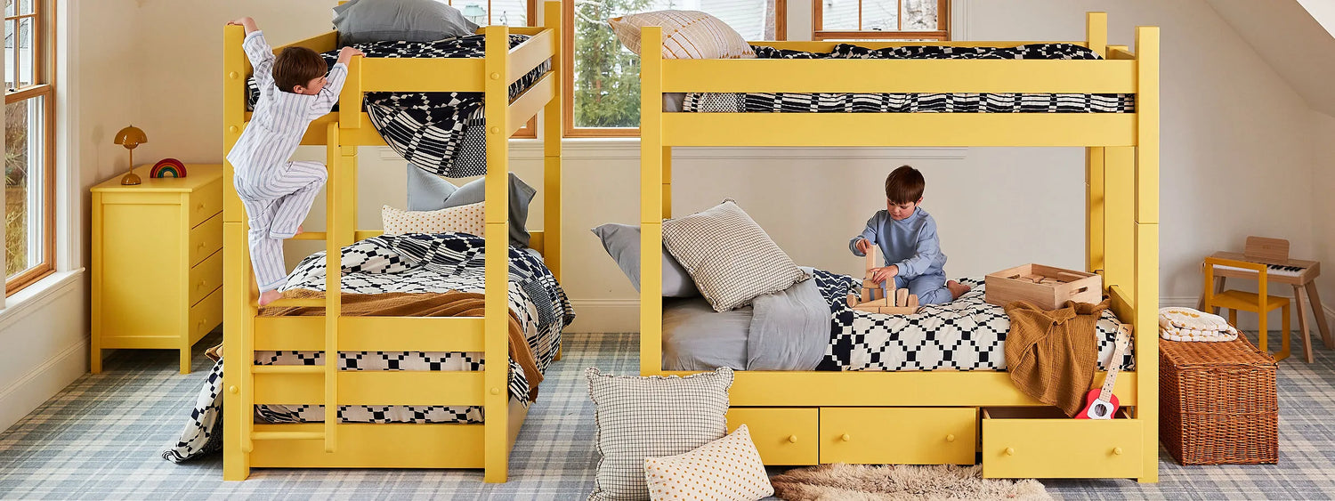 Two bright yellow twin over twin bunk beds in an attic bedroom. A small boy walks up the ladder of one bunk bed, while another small boy plays with wooden blocks on the bottom bunk of the other bunk bed.