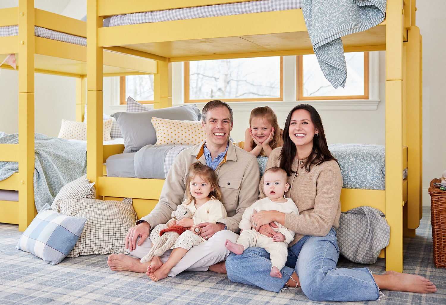 A happy and smiling family of five sits and poses on the floor in front of a bright yellow bunk bed with light blue bedding in various patterns.